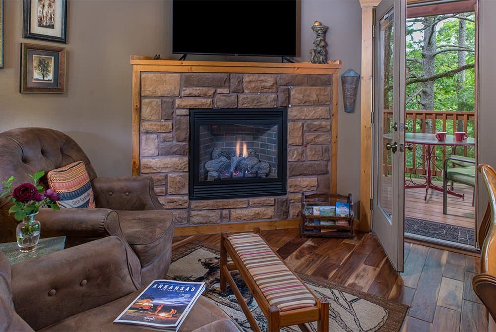 Cozy cabin interior featuring a large stone fireplace with a mounted flat-screen TV, comfortable leather armchairs, and an open door leading to a scenic wooded balcony.