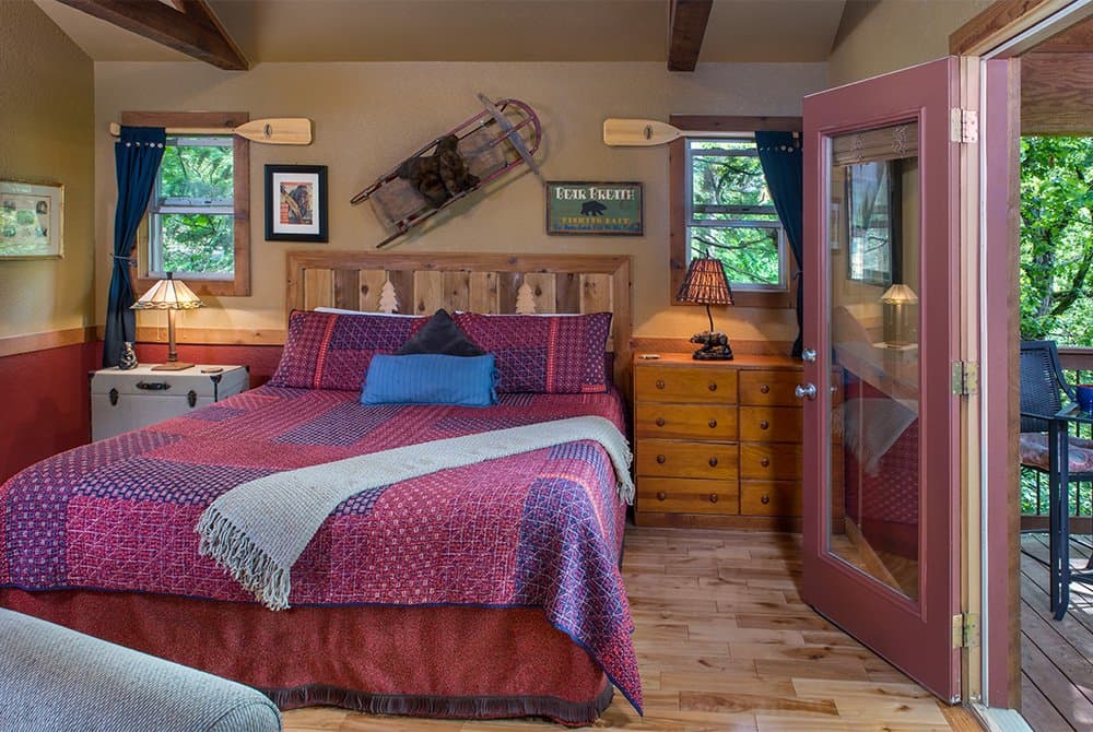 Bedroom interior with a king bed featuring a colorful purple and red quilt, rustic wall decor including oars and a sled, and an open door leading to a balcony.