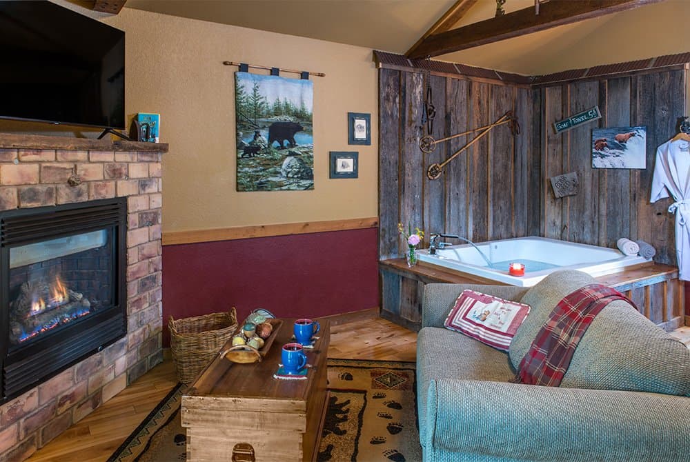 Cozy cabin interior featuring a brick fireplace with a roaring fire, a comfortable armchair, and an indoor Jacuzzi tub nestled in a corner with rustic wood paneling.