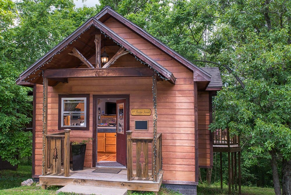 Exterior view of a rustic wooden cabin with a gabled porch and decorative branch-style trusses, surrounded by lush green trees.