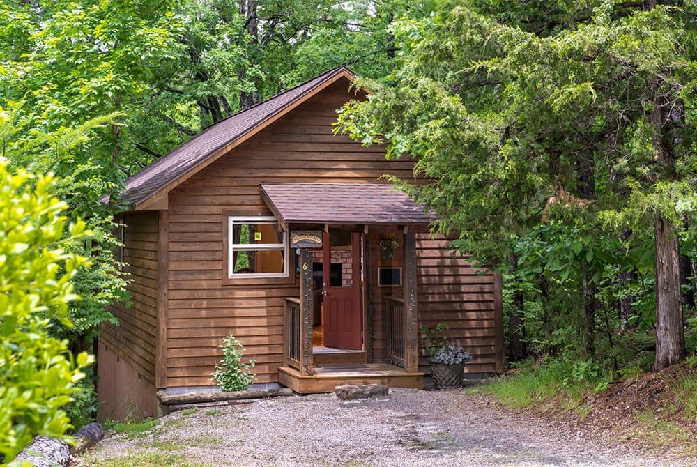 A rustic wooden cabin with a small covered porch and entrance, nestled among lush green trees and dense forest foliage.