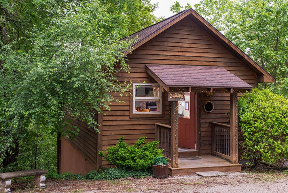 Exterior view of a small, rustic wooden cabin with a gabled roof and a covered entryway, tucked into a lush, green forest area.