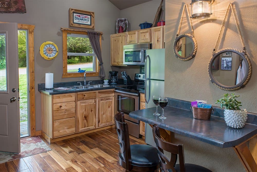 A modern cabin kitchen featuring light wood cabinetry, stainless steel appliances, and a gray breakfast bar with two wooden chairs and wine glasses.