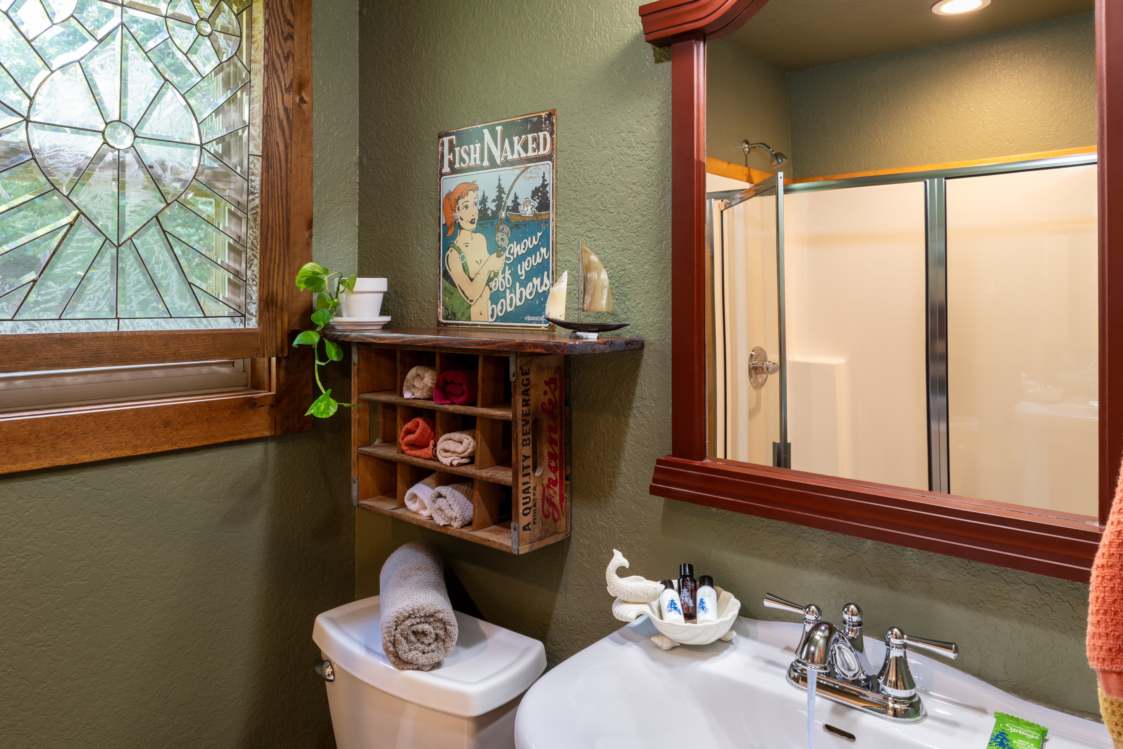 A rustic bathroom featuring olive green walls, a wooden shelf with rolled towels, a stained-glass window, a white sink, and a shower reflected in a wood-framed mirror.