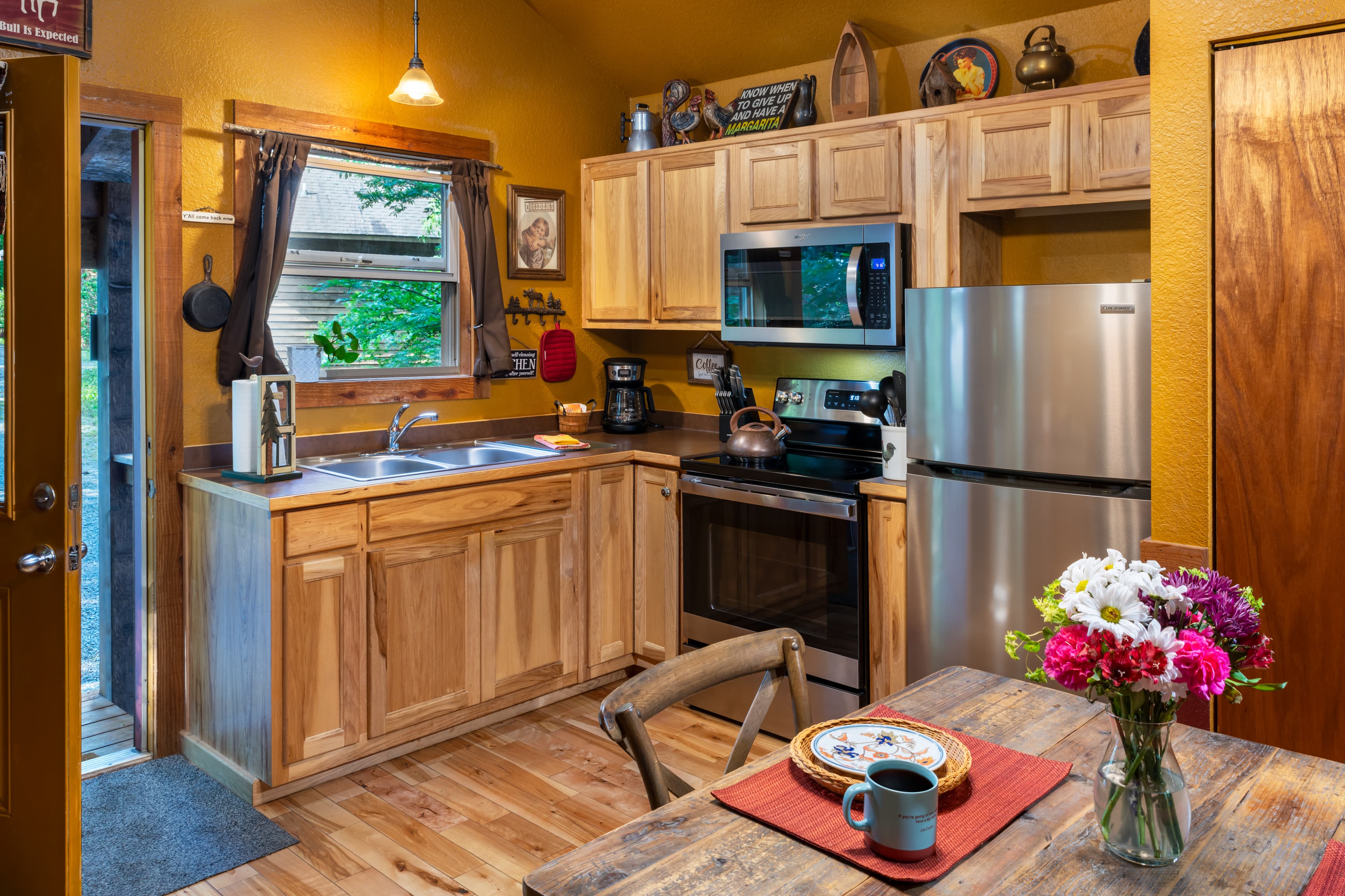 A warm, wood-accented kitchen in a rustic cabin featuring light wood cabinets, stainless steel appliances, and a wooden dining table with a fresh bouquet of flowers.