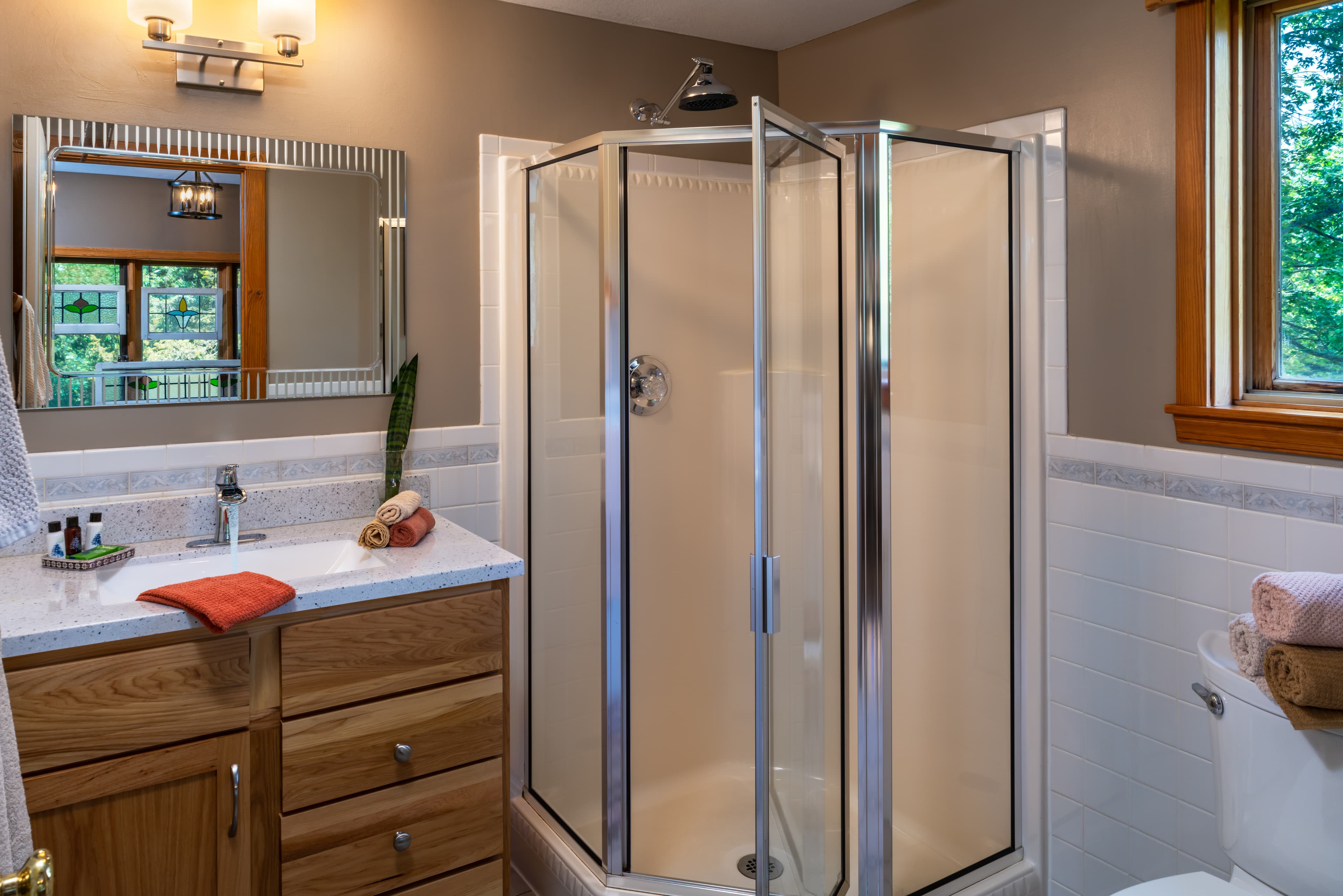 A clean, modern cabin bathroom featuring a corner glass shower stall, a light wood vanity with a white countertop, and a large mirror reflecting a window with stained-glass accents.
