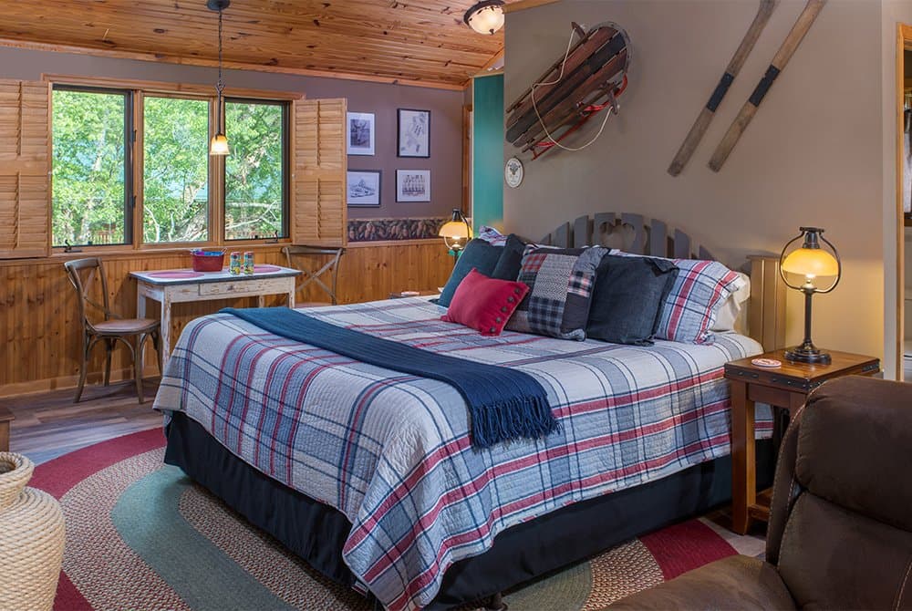 A cozy rustic bedroom featuring a king-sized bed with a blue and red plaid comforter, wood-paneled walls, and nautical-themed decor including wooden oars and a sled mounted above the headboard.