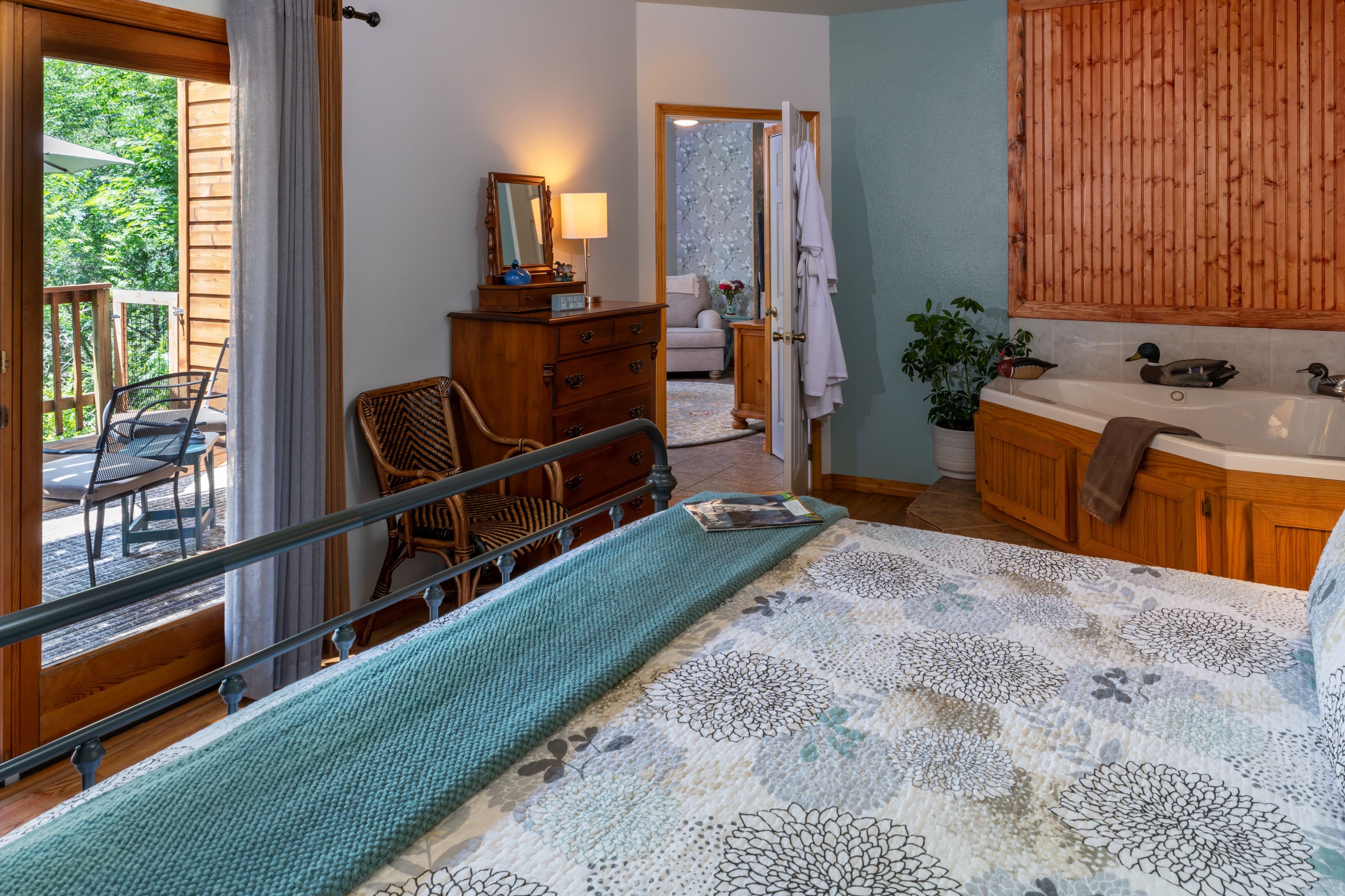 A cozy bedroom featuring a floral print bedspread, a wooden dresser, and a Jacuzzi, with a glass door opening onto a private wooden balcony.