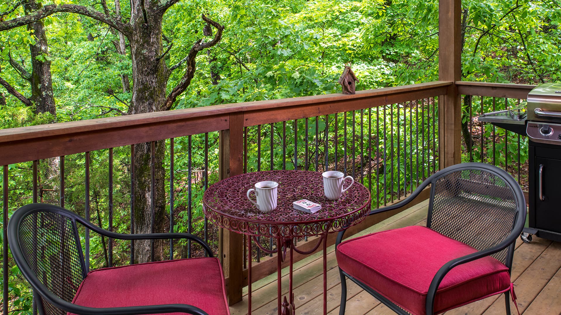 A cozy balcony with two red cushioned chairs, a small table, and lush greenery in the background.