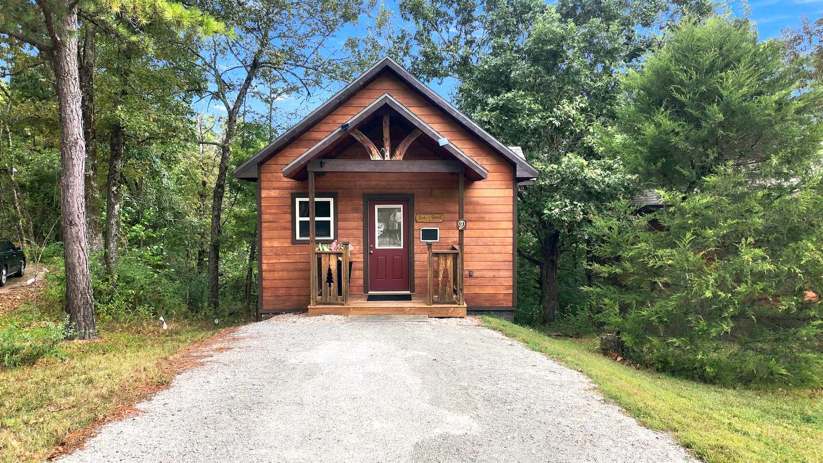 A small wooden cabin with a red door, surrounded by greenery, sits at the end of a gravel path.