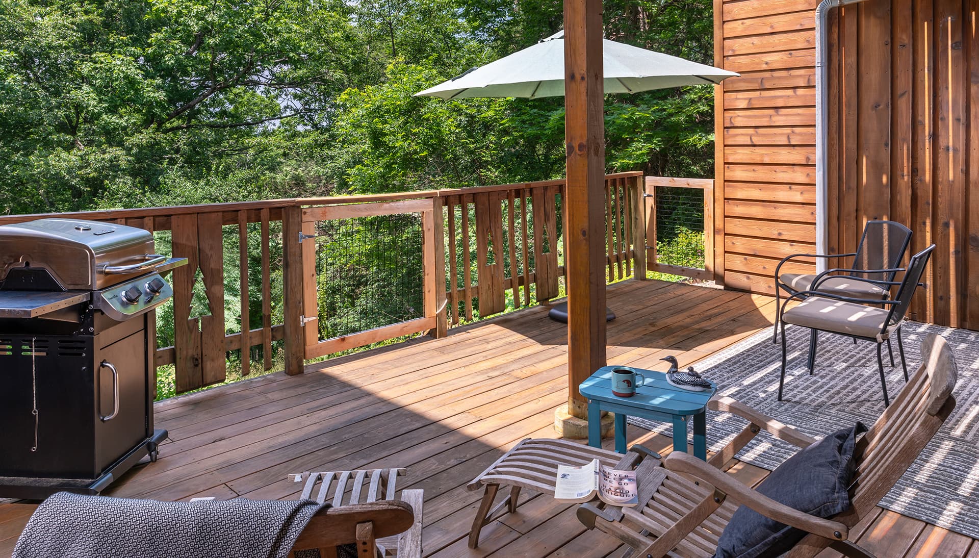 A wooden deck with a grill, chairs, a small table, and an umbrella surrounded by greenery.