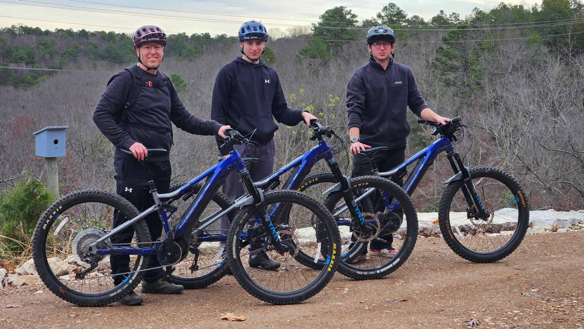 Three mountain bikers pose with their bikes on a dirt path surrounded by trees.