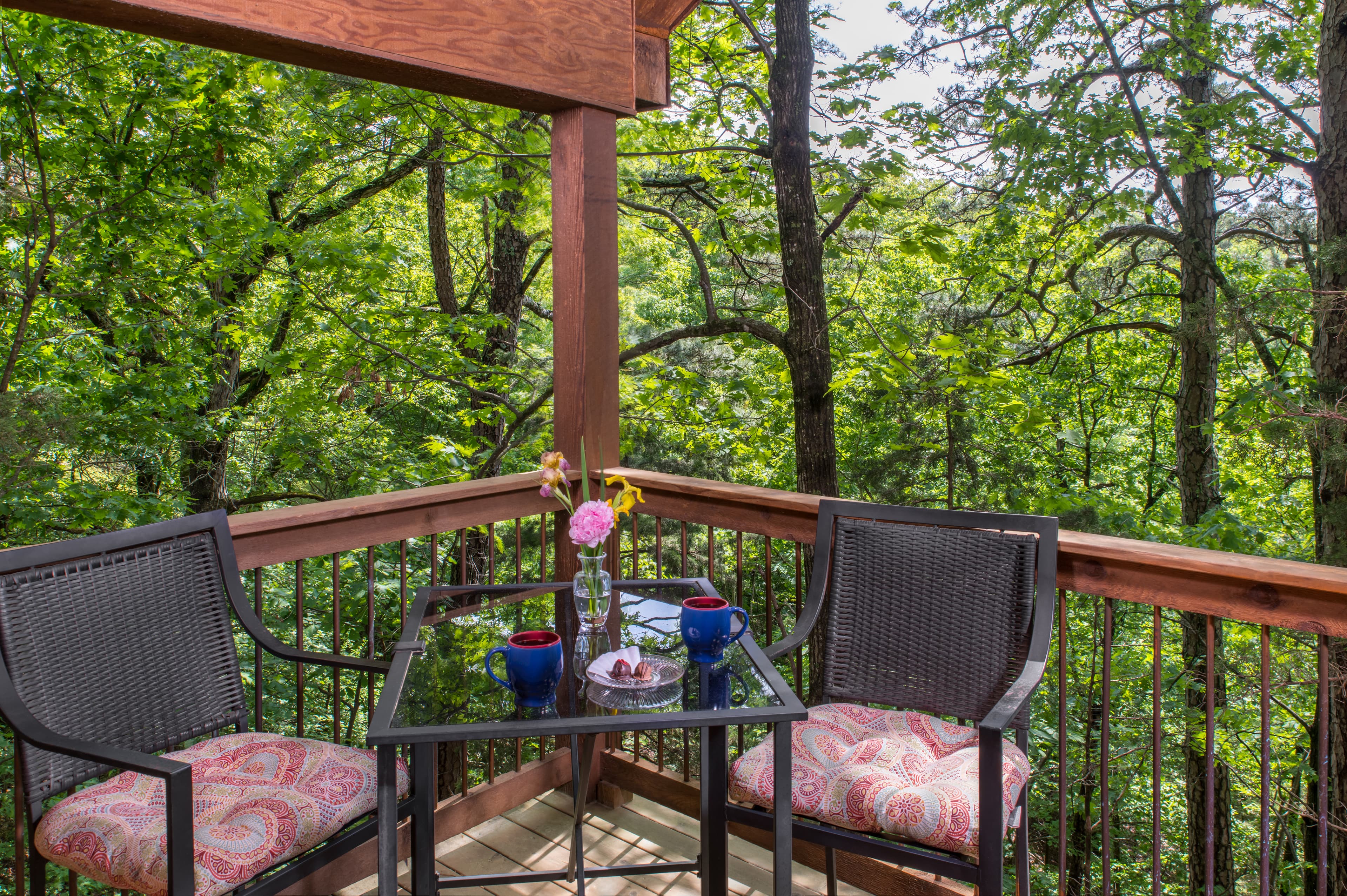 A cozy balcony with two chairs, a table, and a view of lush green trees.