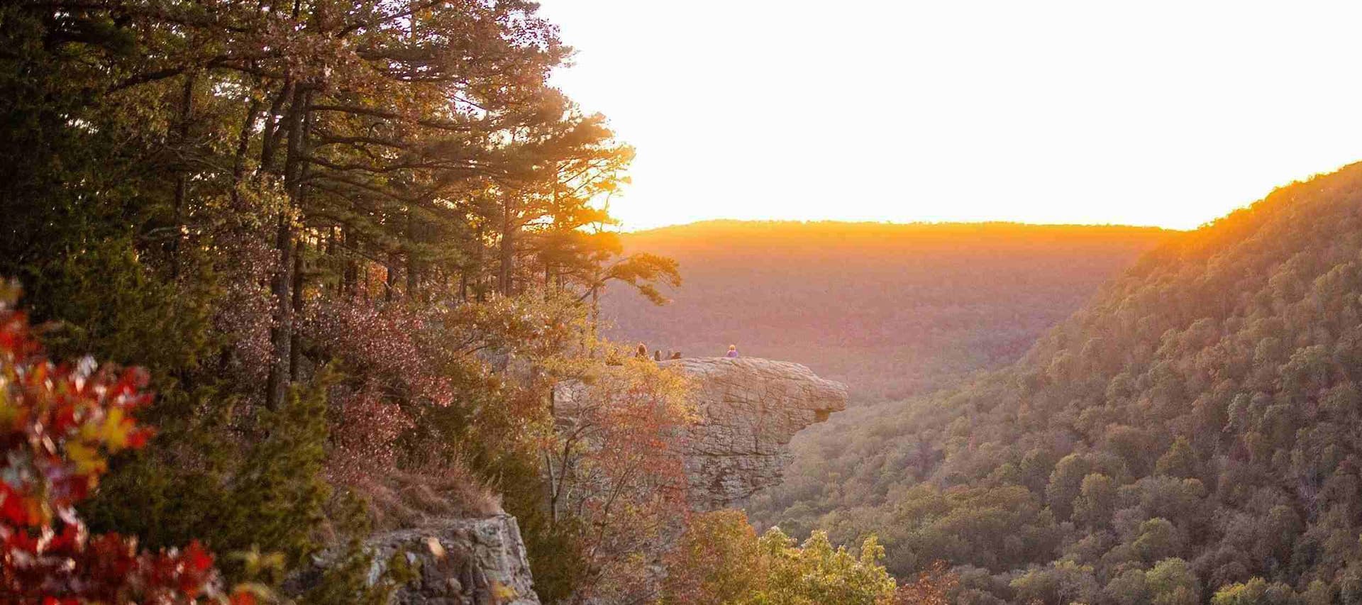 Sunset view over a serene valley from a rocky ledge surrounded by trees.