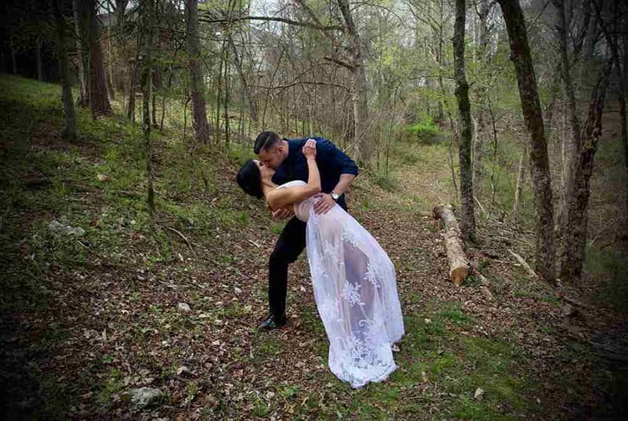 A couple shares a romantic kiss in a forested area, with one partner in a flowing white dress.